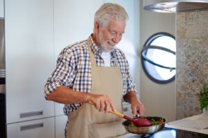what are ADLs Smiling bearded senior man cooking vegetables holding a frying pan. People in the kitchen preparing dinner. An elderly man alone cooking at home