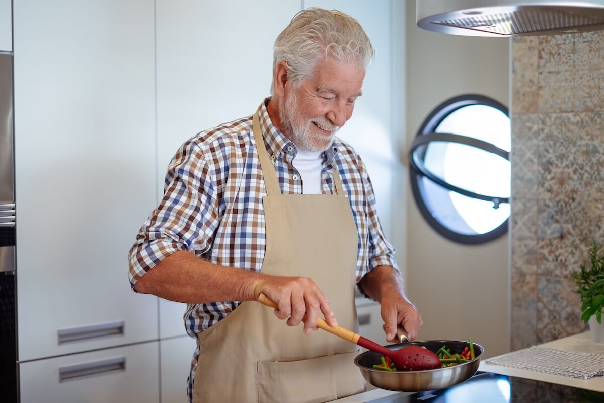 what are ADLs Smiling bearded senior man cooking vegetables holding a frying pan. People in the kitchen preparing dinner. An elderly man alone cooking at home