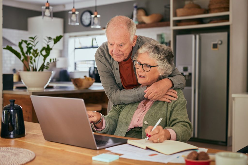 Elderly couple collaborating on tasks with a laptop and notebook at home while sharing an affectionate moment. Old man and elderly woman managing documents and finances together. Mature woman working from home while husband embracing her from behind.
