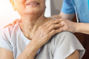 A senior woman touches the hand of a caregiver and smiles over her shoulder, representing memory care safety.